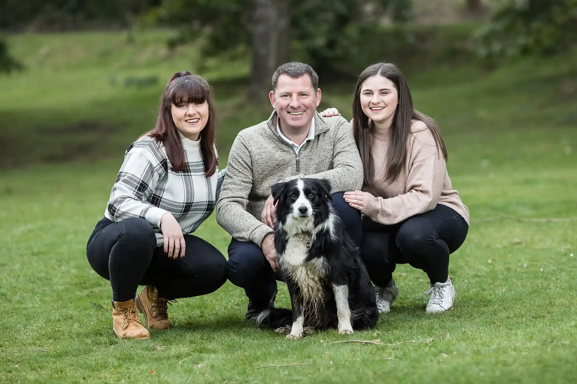 Three people squat beside a black and white dog outdoors on a grassy area with trees in the background.