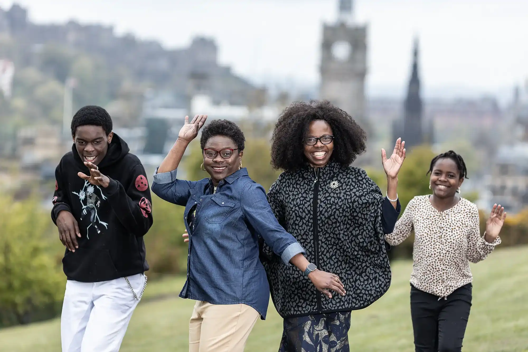 Four people are outdoors in a park, posing and smiling in a line. A clock tower is visible in the background.
