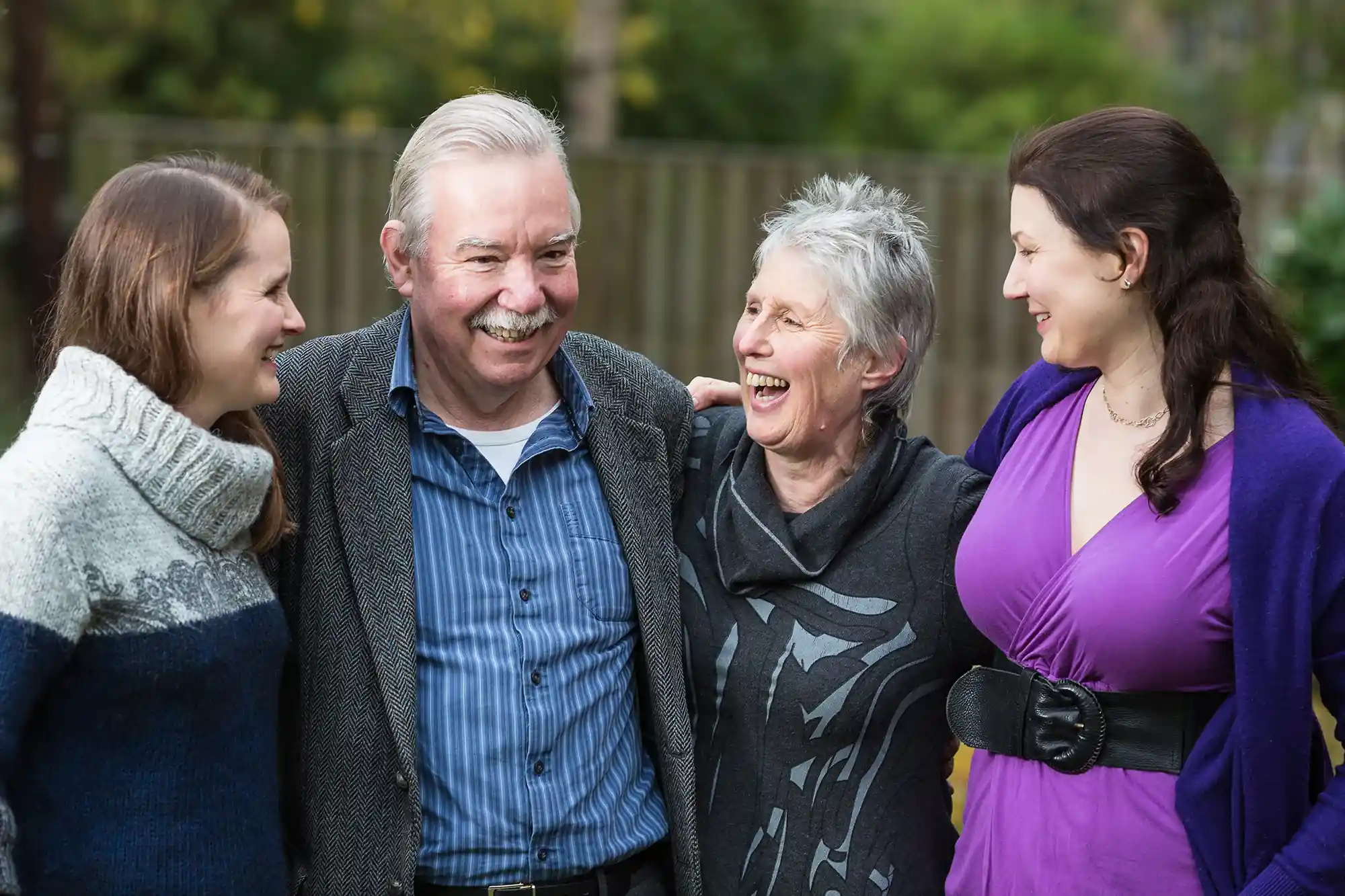 Four people are posing outside, smiling and looking at each other. The group includes two women on the left, a man in the middle, and another woman on the right. They appear to be enjoying a pleasant moment together.