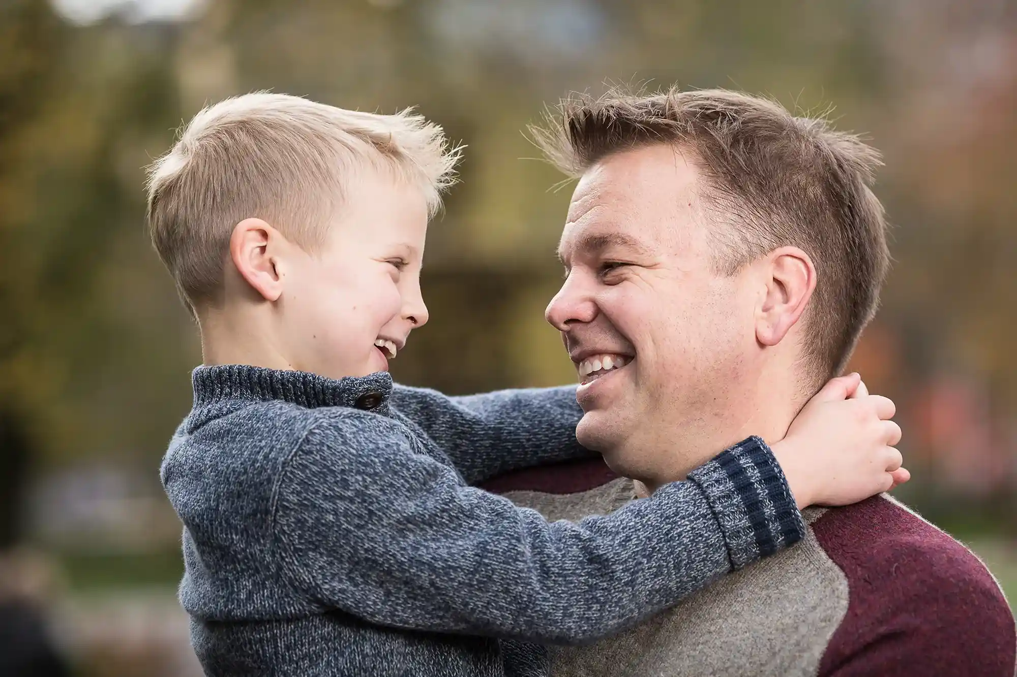 A man holds a smiling child who has their arms around his neck. Both are looking at each other joyfully. The background is blurred and appears to be outdoors.