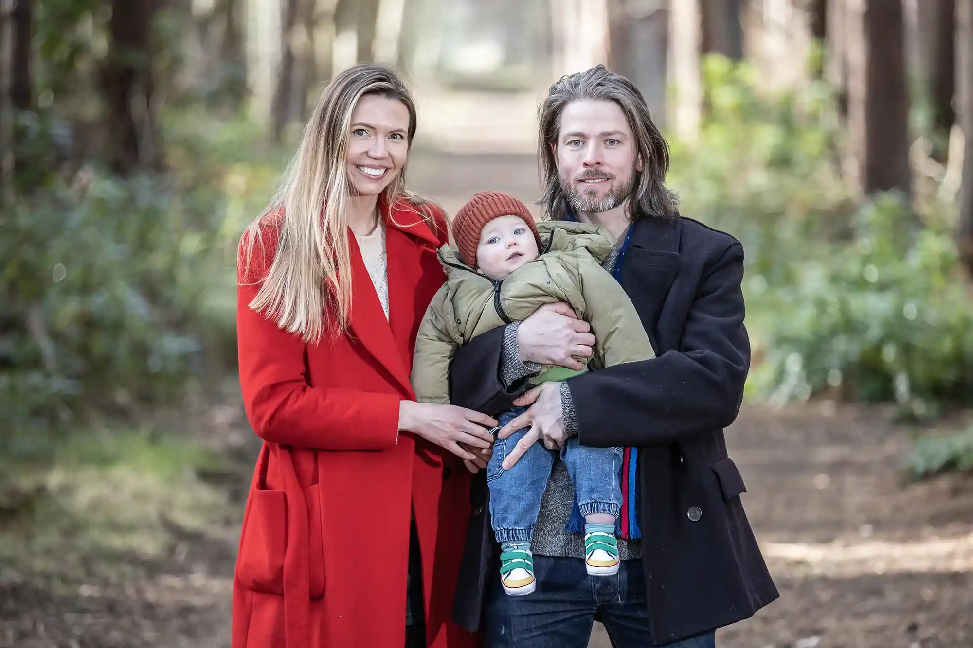 A couple stands in a forest path, with the man holding a baby dressed in winter clothes. The woman is wearing a red coat, and the man is wearing a dark coat.