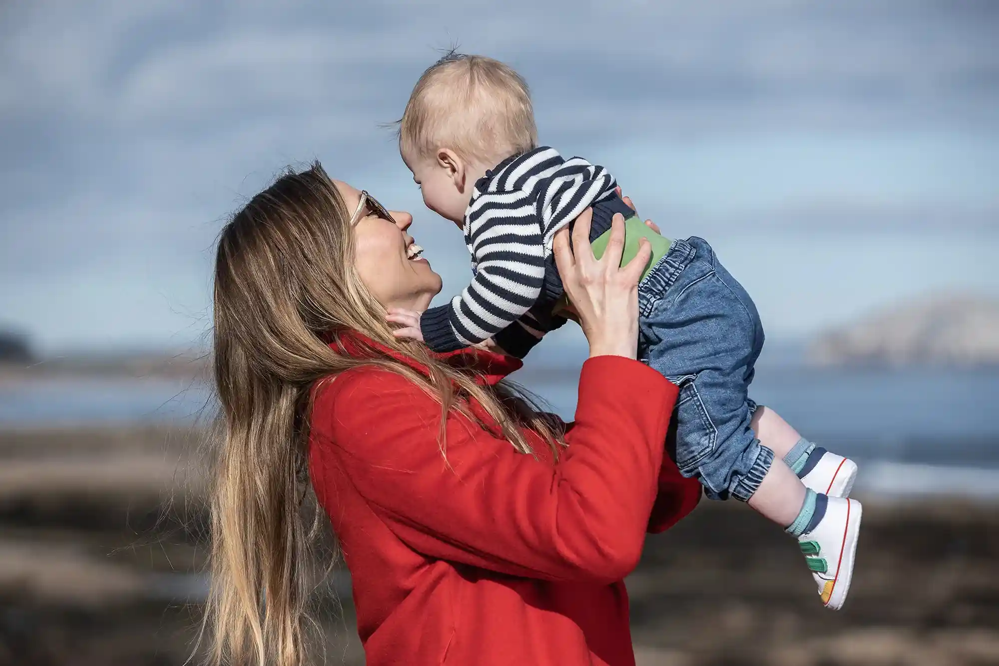Woman in a red coat holds a baby wearing a striped shirt and blue pants. Both are outdoors on a cloudy day near water.