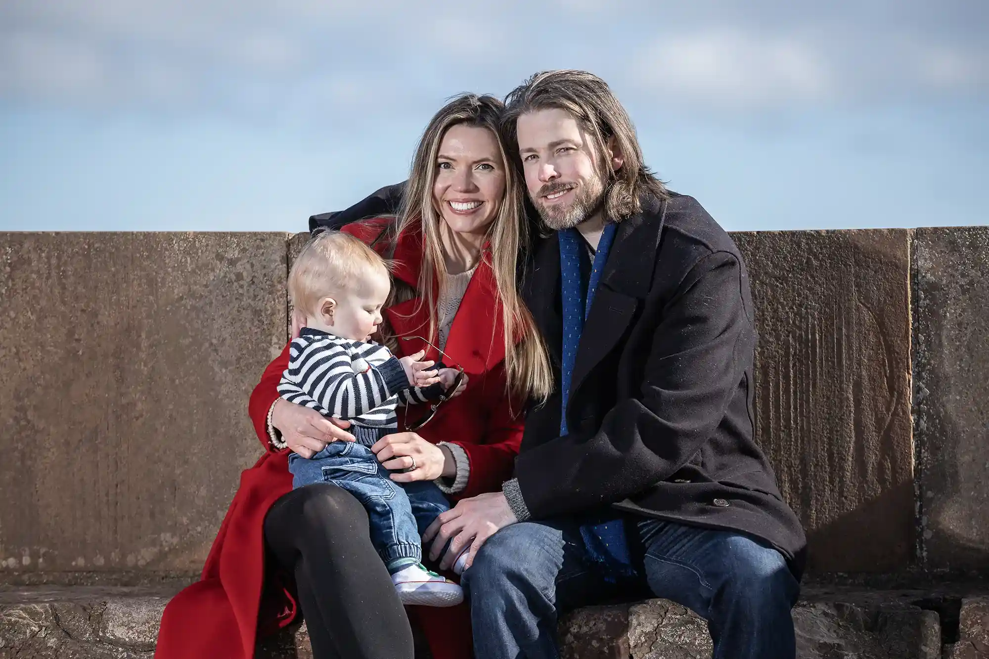 A smiling woman wearing a red coat is seated with a man in a black coat. They are holding a baby in a striped shirt and blue jeans, seated on a stone bench with a cloudy sky in the background.