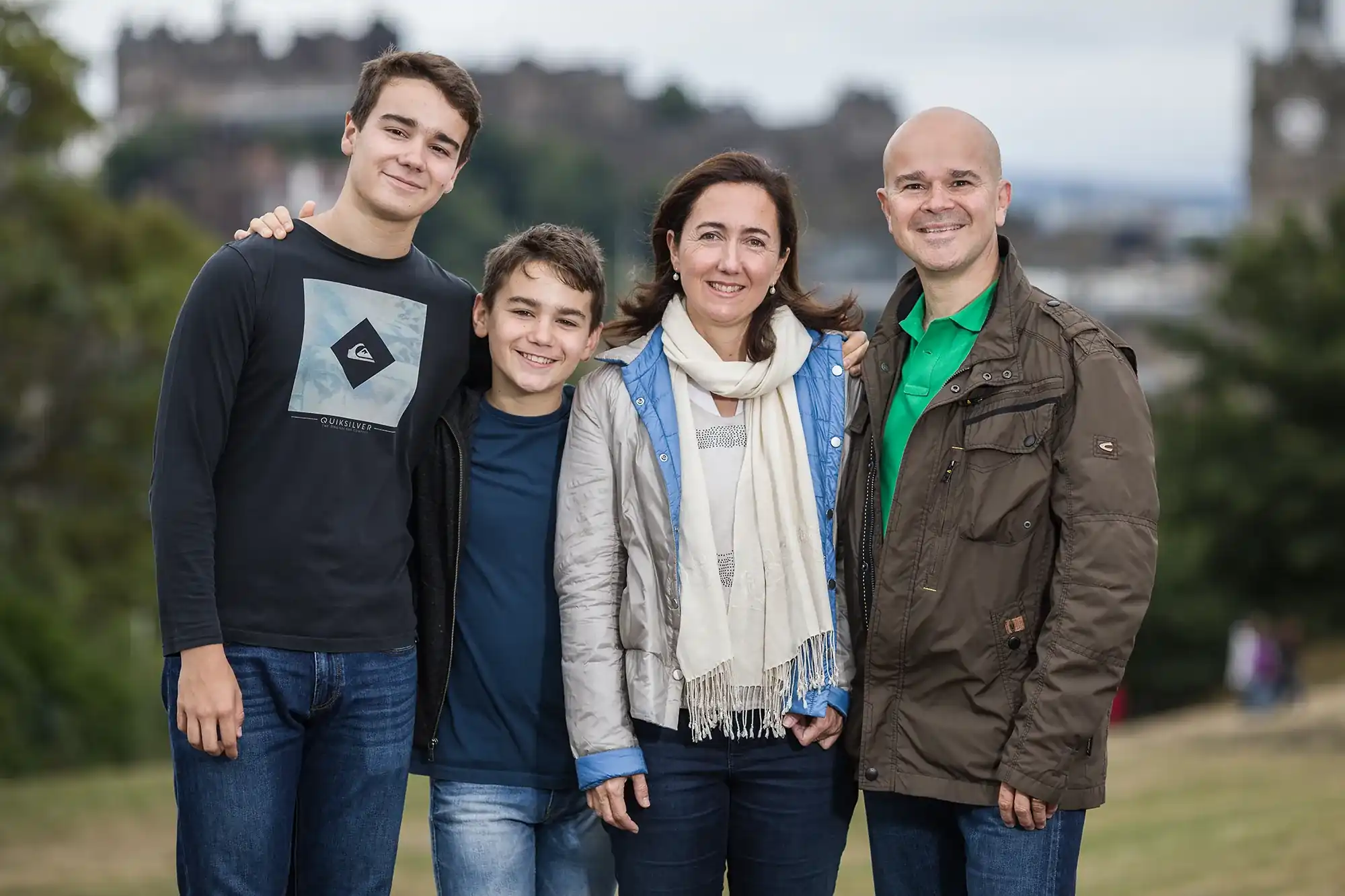 Four people, consisting of two adults and two boys, stands close together outdoors with an overcast sky and blurred trees and buildings in the background.