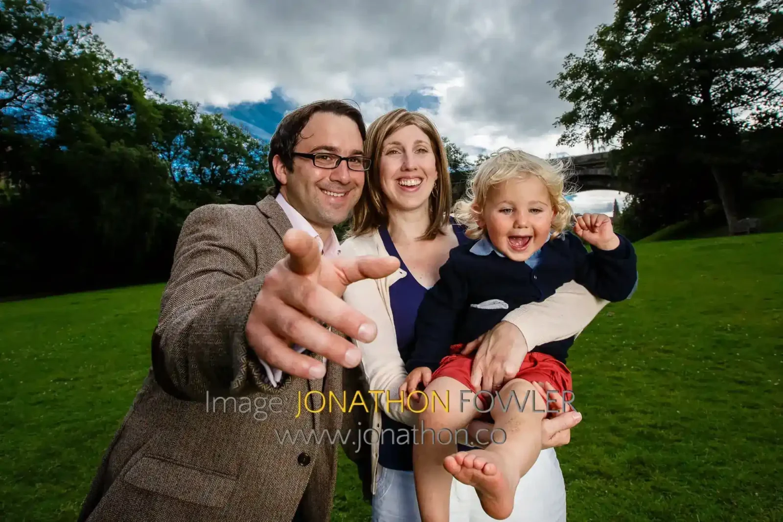 A smiling man points at the camera while standing next to a smiling woman holding a laughing young child in her arms. They are outdoors on a grassy field with trees and a cloudy blue sky in the background.