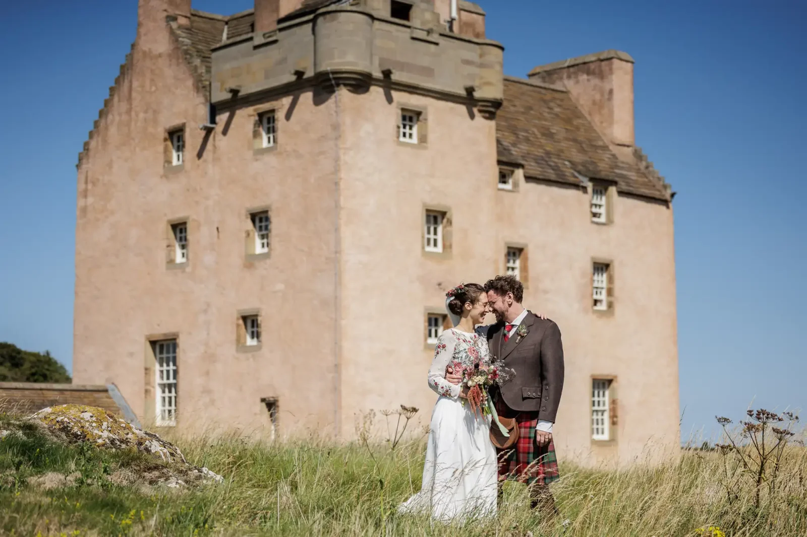 A bride in a white dress and groom in a kilt stand in tall grass, embracing and smiling, holding a bouquet. Behind them is a large, pinkish stone castle with small windows, set against a clear blue sky on a sunny day.