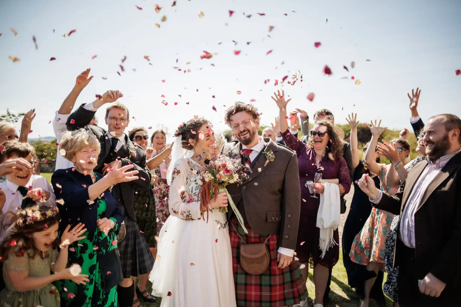 Fenton Tower wedding photographer photo of a bride and groom, both smiling, stand together as guests surround them outdoors, tossing flower petals in celebration. The groom wears a kilt, and the bride holds a bouquet. The crowd is joyful, with hands raised and confetti petals floating in the air.
