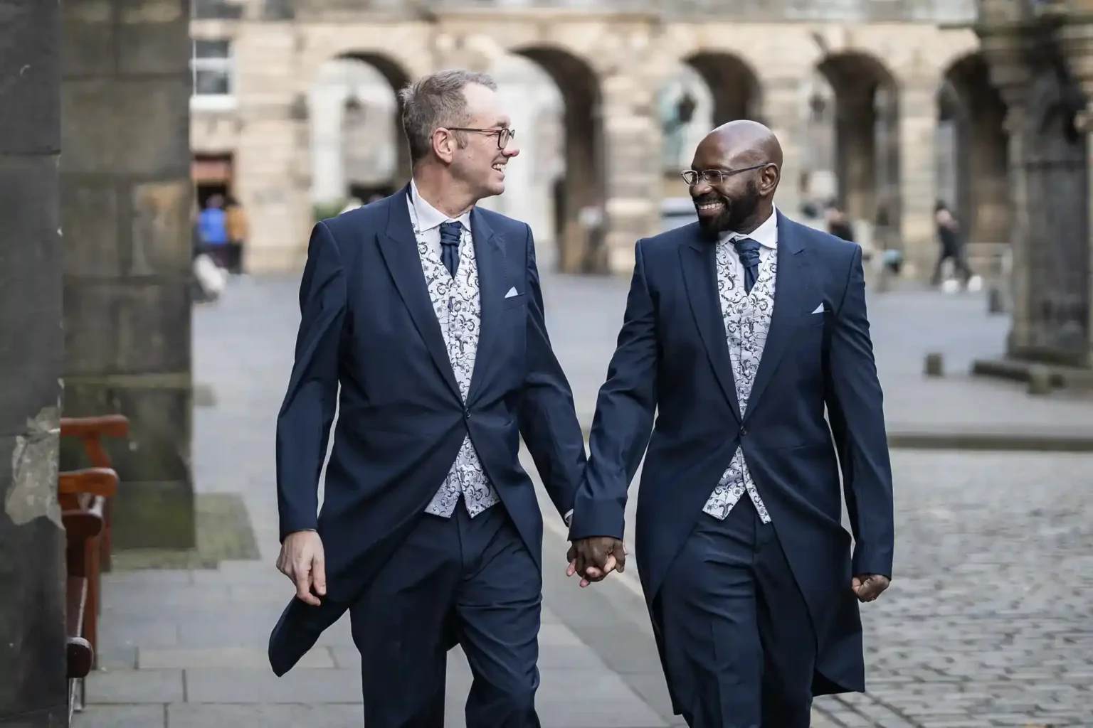 Two men dressed in matching navy blue tailcoats, patterned waistcoats, and ties walk hand in hand down a cobbled street, smiling warmly at each other, with historic stone buildings in the background.