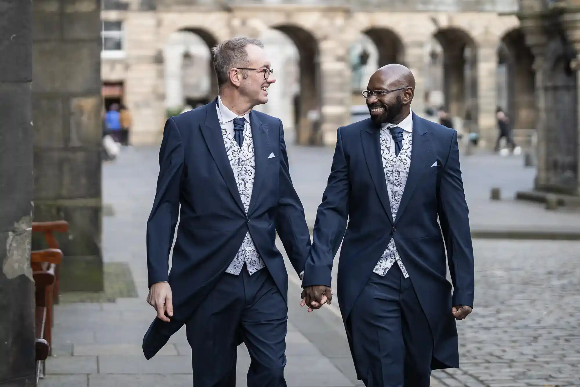 Two men dressed in matching navy blue tailcoats, patterned waistcoats, and ties walk hand in hand down a cobbled street, smiling warmly at each other, with historic stone buildings in the background.