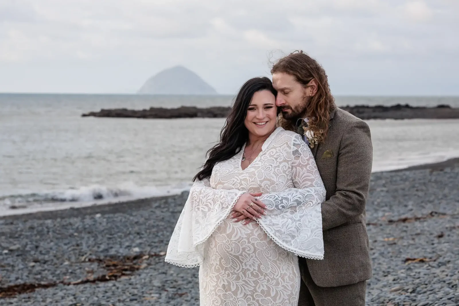 A smiling pregnant woman in a white lace dress stands on a rocky beach, her hands on her belly. A man in a brown suit hugs her from behind, resting his chin on her head. The sea and a distant island are visible under a cloudy sky.