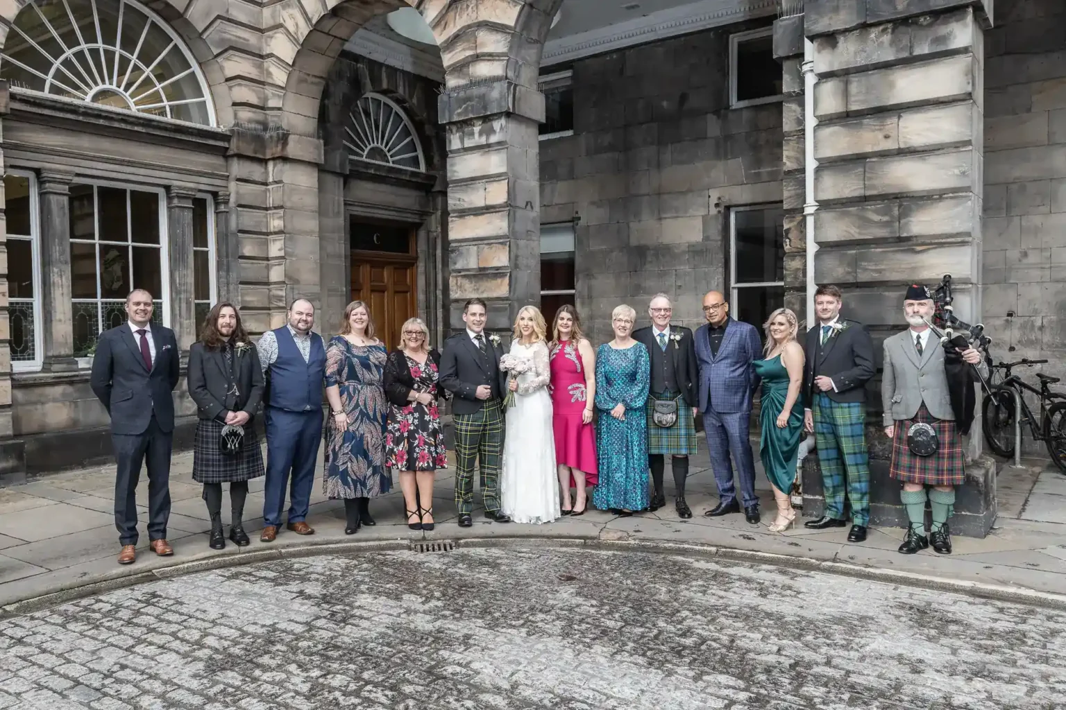A wedding group poses outside a historic stone building. The bride in white and groom in a kilt stand centre, surrounded by family and friends in formal attire, some wearing kilts. The mood is celebratory and joyful, with arched doorways and windows as the backdrop.