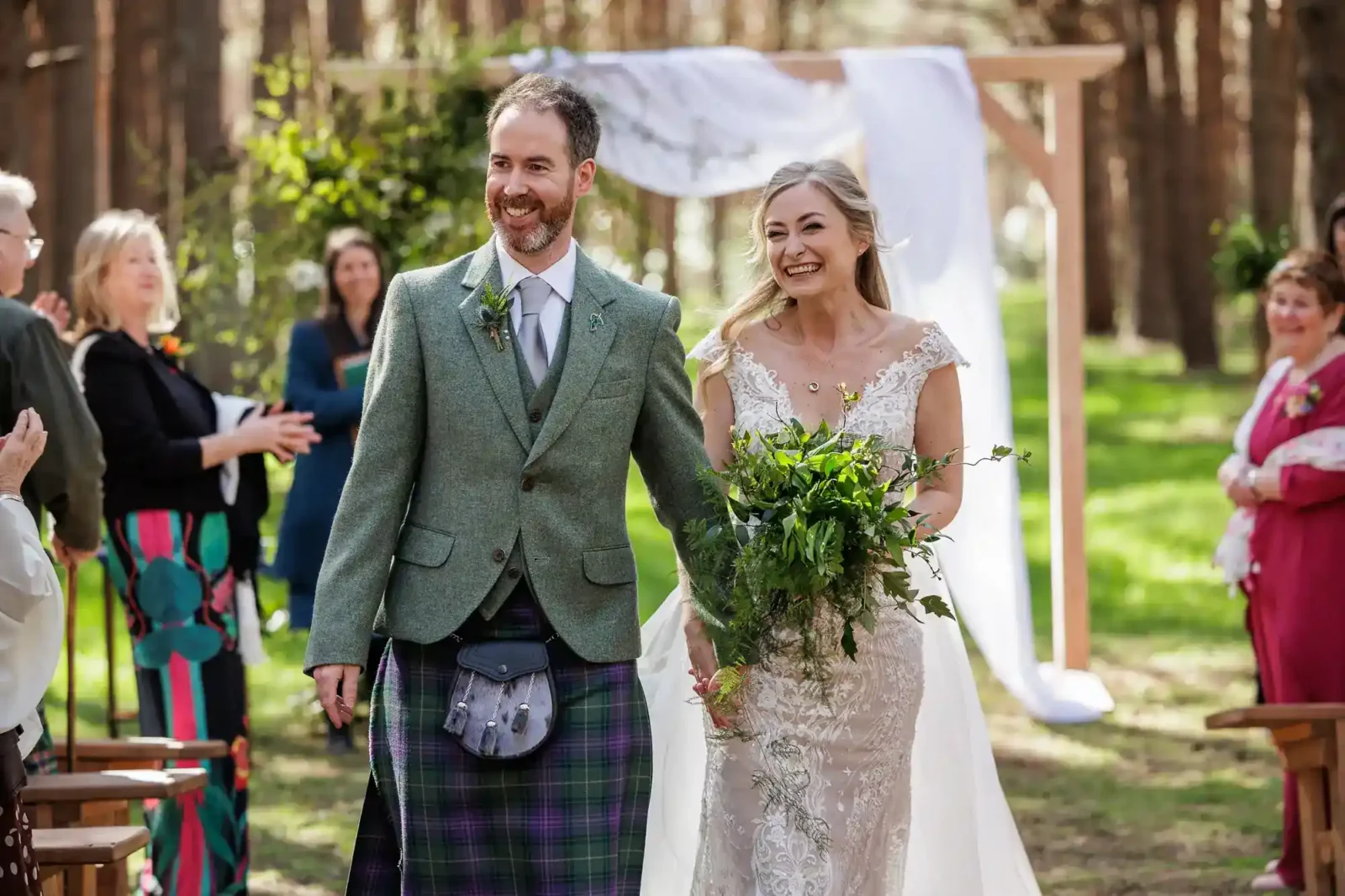 A smiling bride in a lace dress and groom in a green jacket and tartan kilt walk hand in hand outdoors. Guests stand and clap, and a wooden wedding arch draped with white fabric is visible behind them in a sunlit forest setting, Harvest Moon.