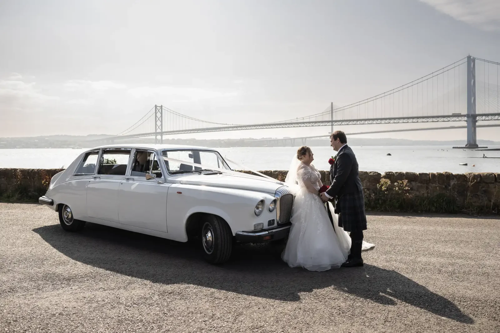 A bride in a white gown and a groom in a traditional Scottish kilt stand holding hands beside a vintage white car, with a large suspension bridge and calm water in the background on a sunny day.