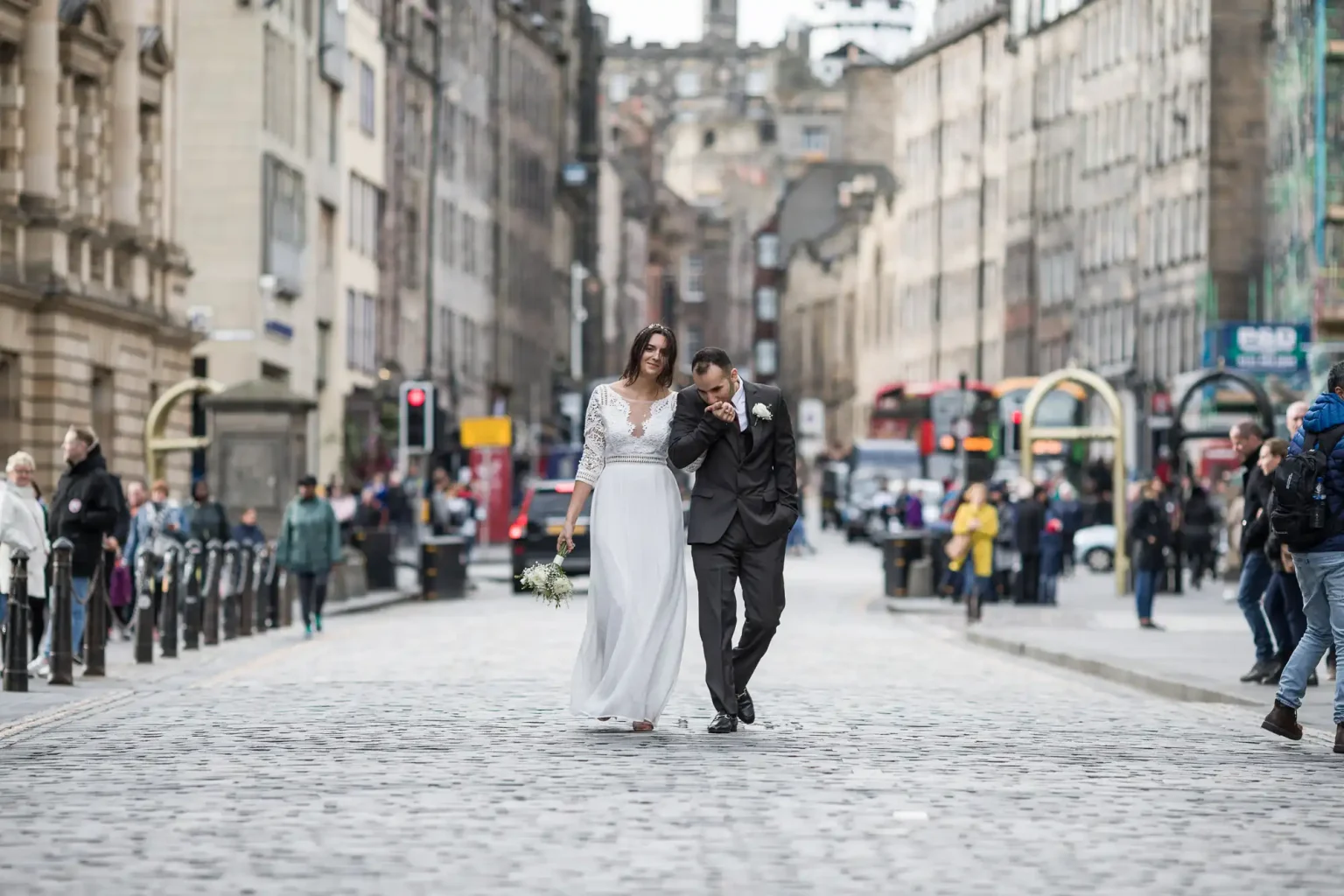 A couple walks arm in arm down a cobblestone street, surrounded by city buildings and pedestrians. The bride is in a white dress holding flowers, and the groom is in a black suit.