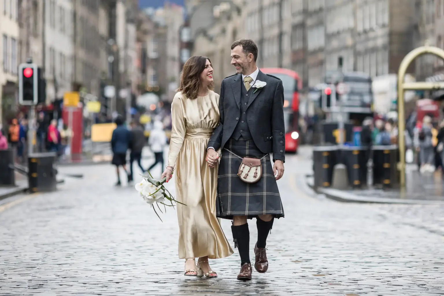 A couple walks hand in hand on a city street; the woman wears a gold dress, and the man wears a kilt and jacket.