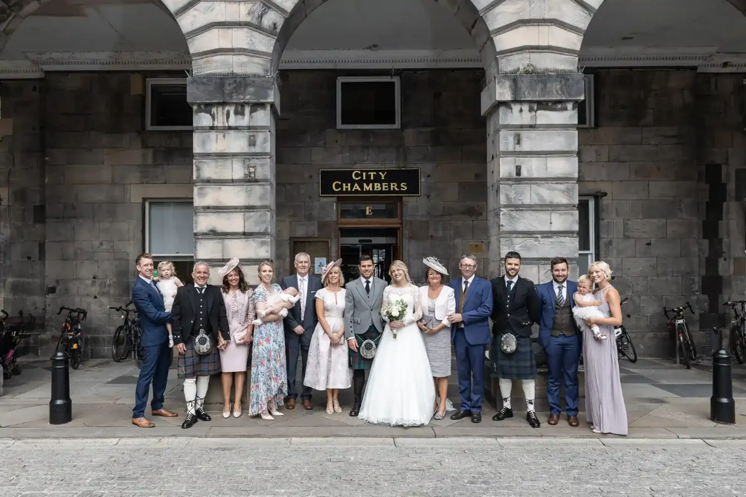 A wedding party of 14 adults and 2 children stands outside a stone building labelled “City Chambers.” The bride is in a white dress, the groom in a kilt. The group is dressed formally, smiling, and holding flowers or children. Bicycles are parked behind them.