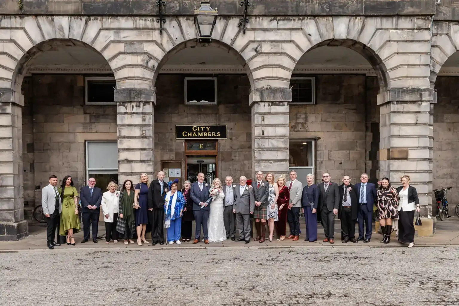A wedding group stands in front of a stone building with three arches marked “City Chambers.” The bride and groom are at the centre in white attire, surrounded by guests in formal clothes. The group is posed in a wide line, smiling for the photo.