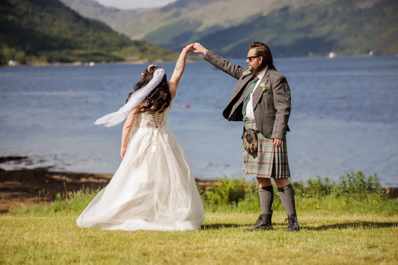 A bride in a white gown and veil spins under the raised hand of a groom in a grey jacket, kilt, and sporran. They stand on grass beside a lake, with green hills and mountains in the background under a clear sky.