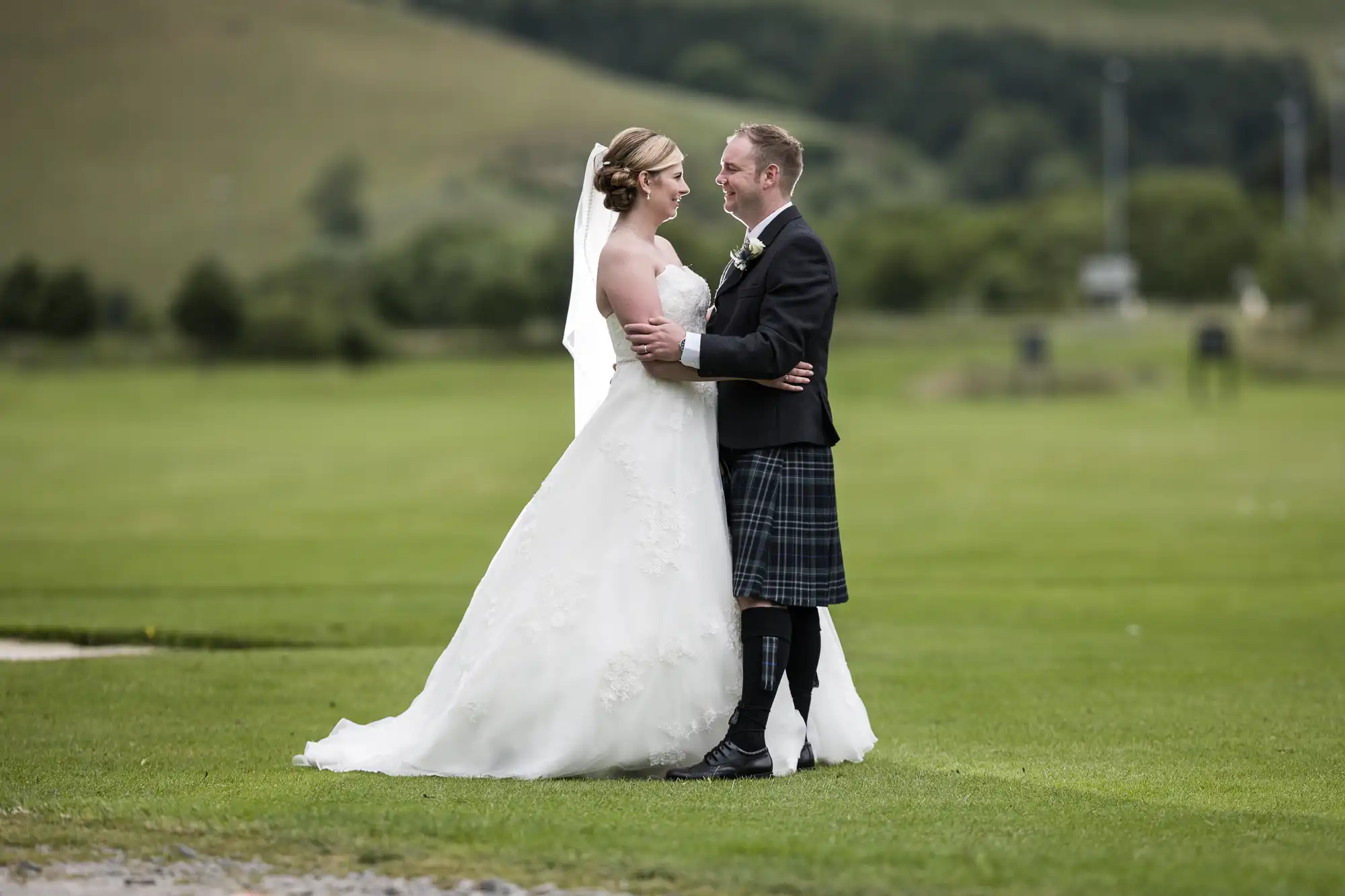 Macdonald Cardrona Hotel wedding photographer, Jonathon Fowler Photography colour image of a bride in a white gown and veil embraces a groom in a black jacket, kilt, and knee-high socks. They stand on green grass with rolling hills and trees in the background, smiling at each other in an outdoor setting.