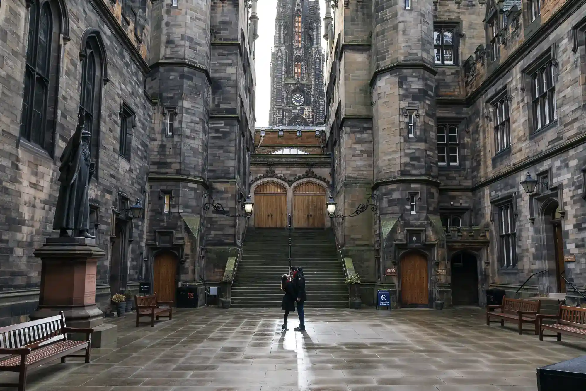 A solitary person stands in a wet, stone courtyard surrounded by tall, old Gothic buildings with arched doors and windows. Stone steps rise to ornate wooden doors, and a statue stands on the left. The scene looks empty, serene, and historical.