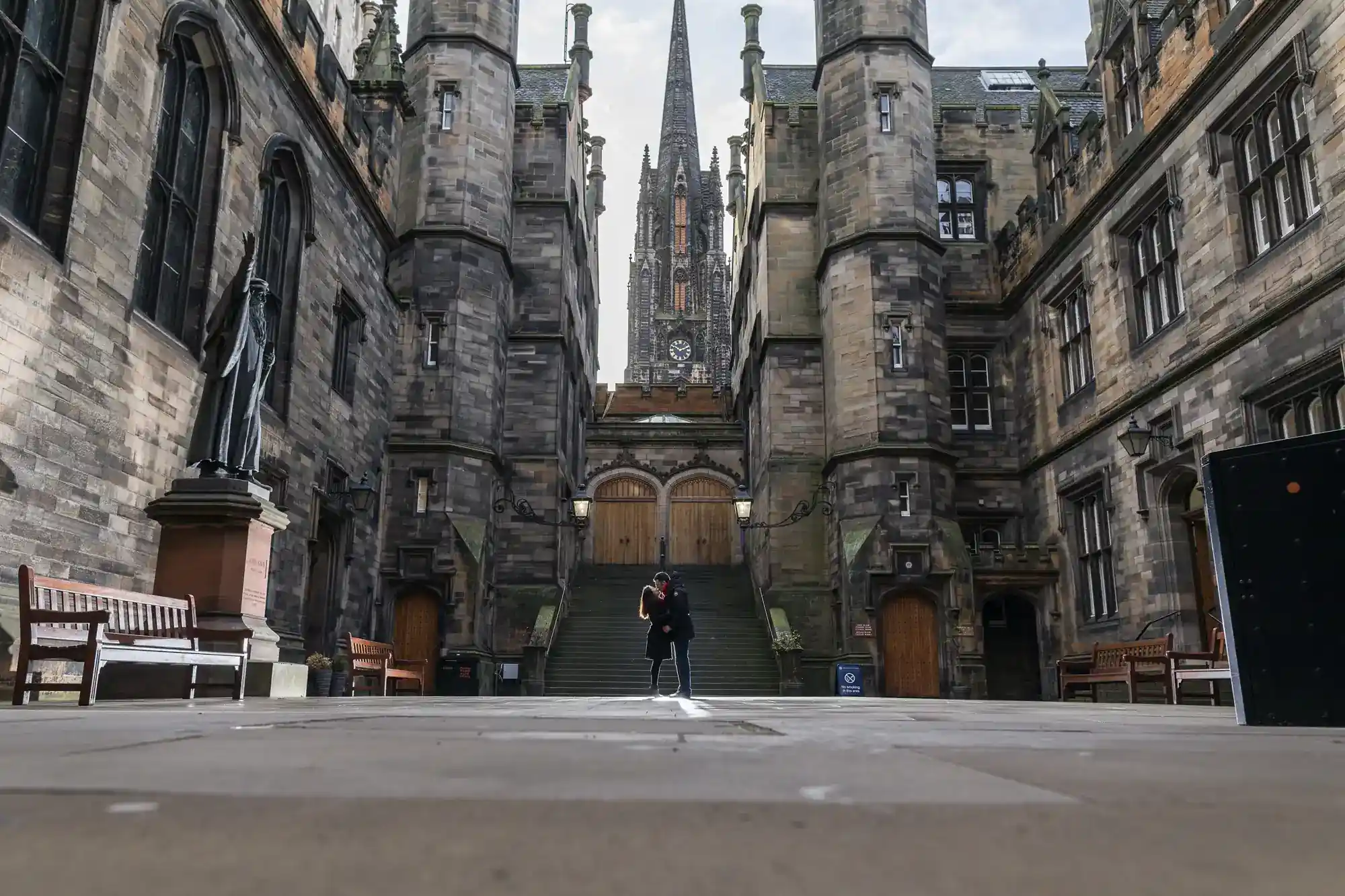 A person stands alone in a large courtyard surrounded by tall, old stone buildings with gothic architecture and arched windows. There are benches, a statue on the left, and a tall clock tower rising in the background. The weather is cloudy.