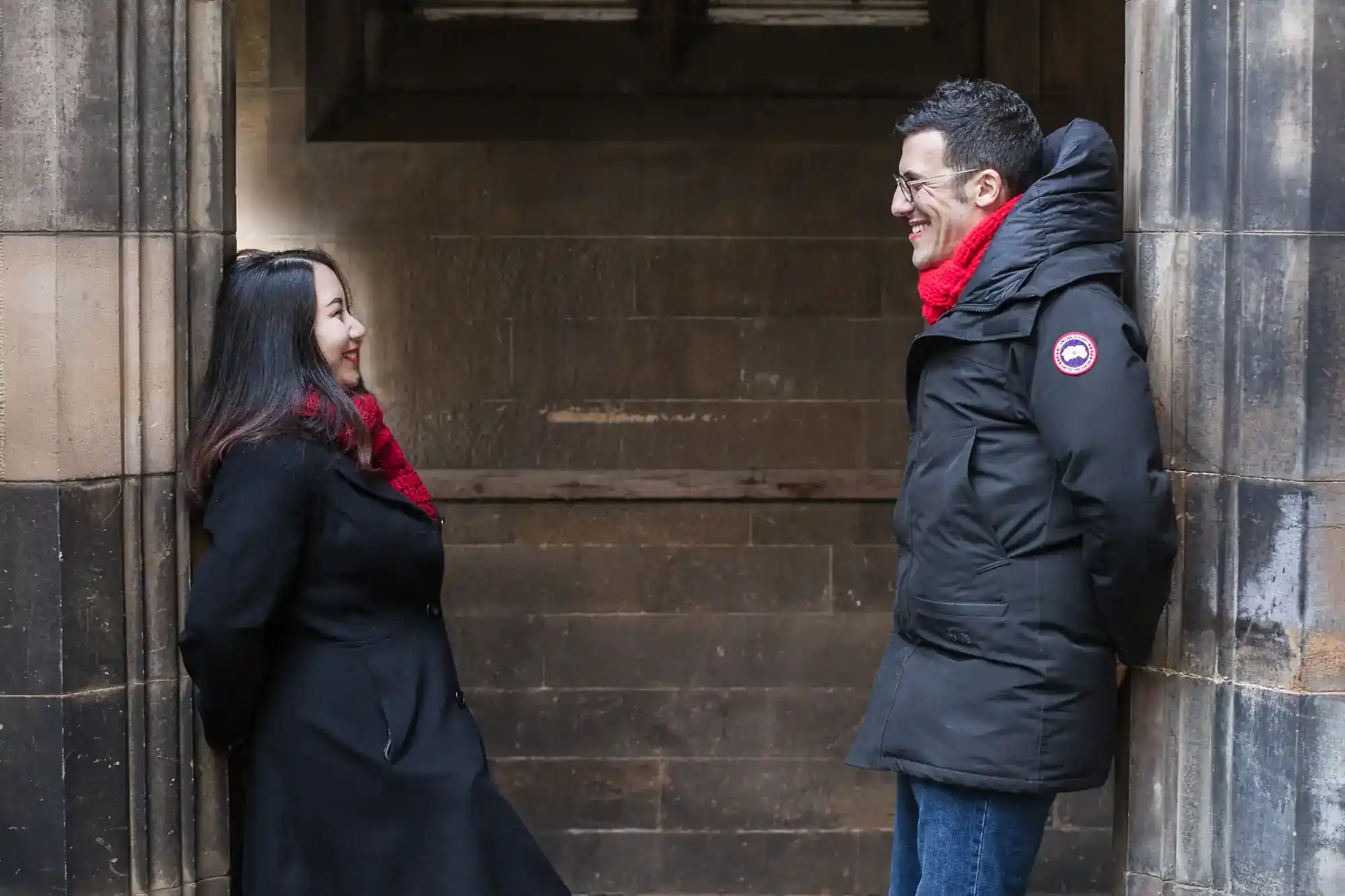 Two people wearing black coats and red scarves stand facing each other, smiling, in a stone archway. The background is a weathered stone wall, and both appear relaxed and happy, suggesting a friendly or warm interaction in a chilly setting.
