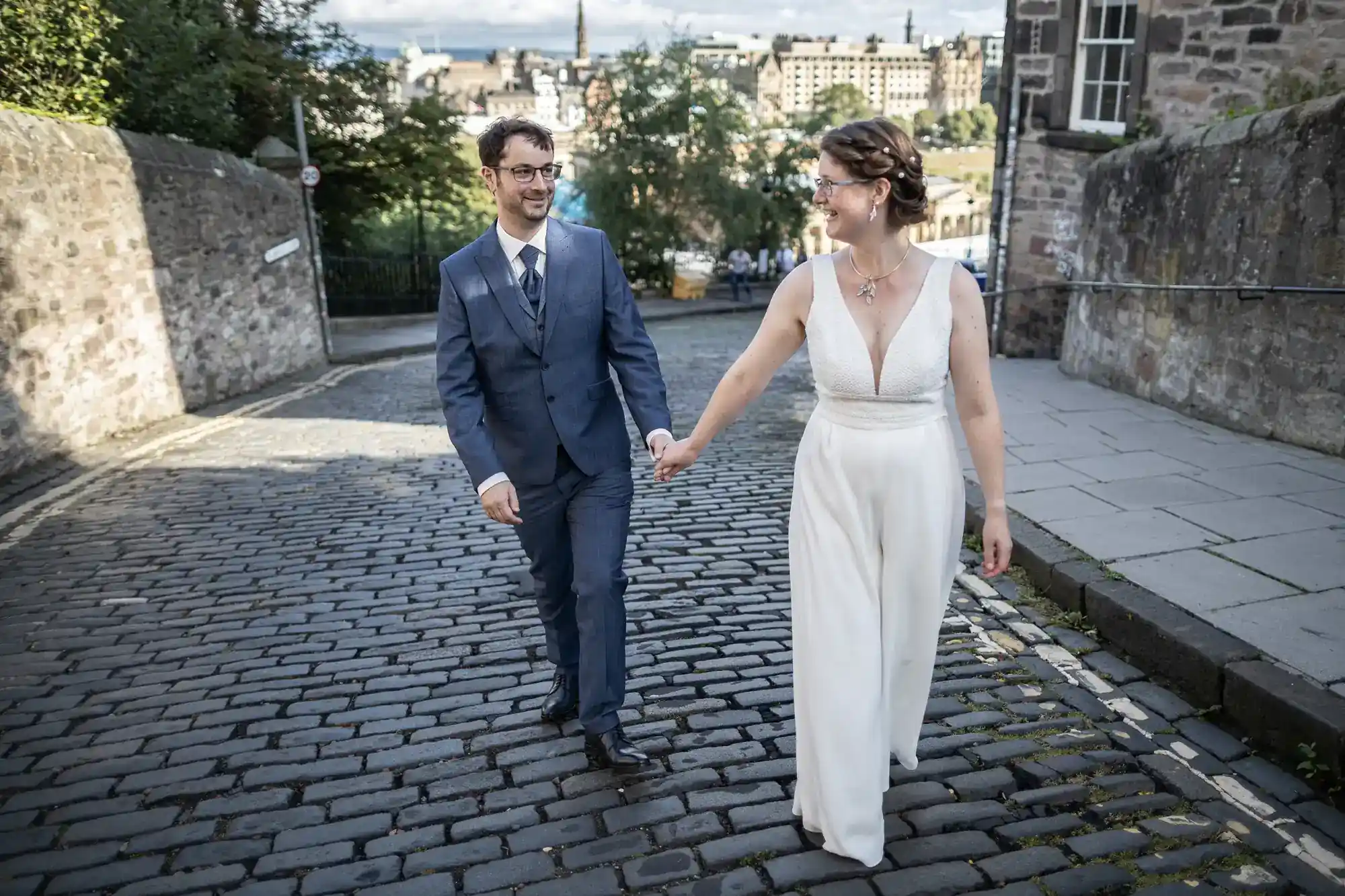 A smiling couple, dressed in a blue suit and a sleeveless white dress, walk hand-in-hand on a cobbled street. Green trees and stone buildings are visible in the background under a partly cloudy sky.