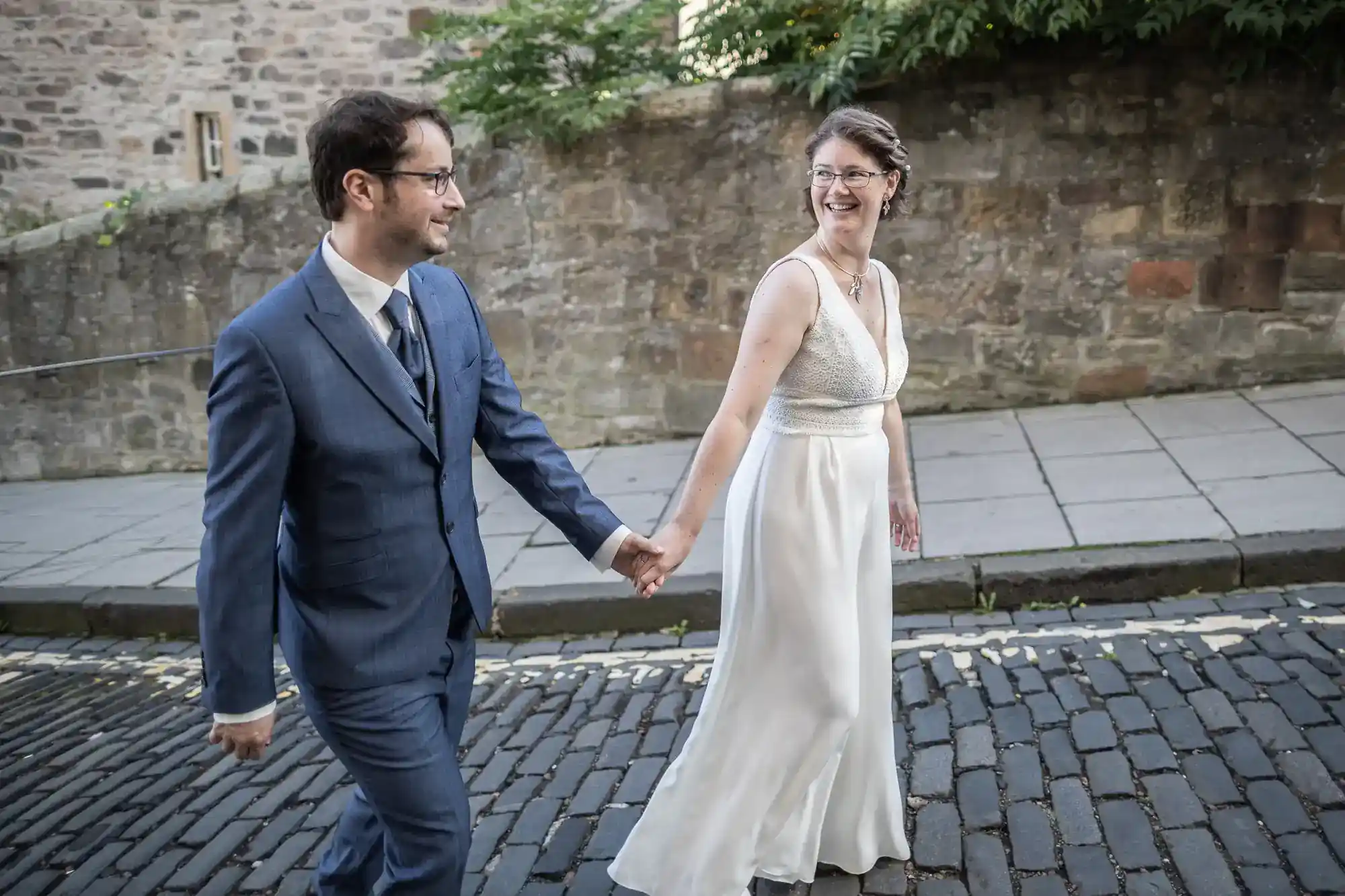 A smiling couple in wedding attire hold hands while walking on a cobbled street. The man wears a blue suit and glasses; the woman wears a sleeveless white dress and glasses. A stone wall and plants are in the background. Both look happy and are looking at each other.