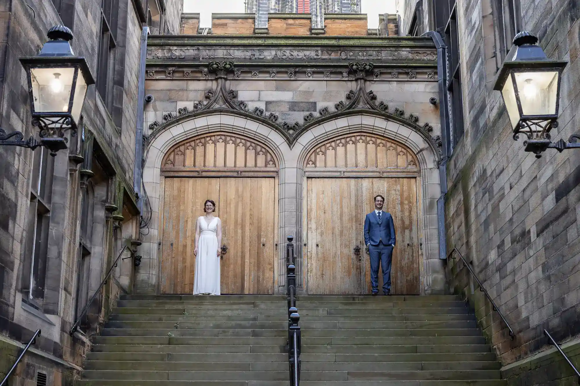 A woman in a white dress and a man in a blue suit stand apart on stone steps, each in front of a large wooden door with arched stone carvings. The setting is a grand, historic stone building with lamps on both sides.