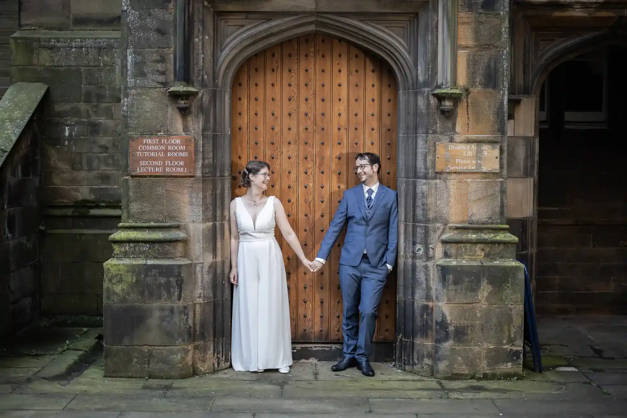 A couple in wedding attire hold hands and smile at each other, standing in front of a large wooden door set in a stone archway. The scene is framed by old stone walls with signs on either side of the doorway.