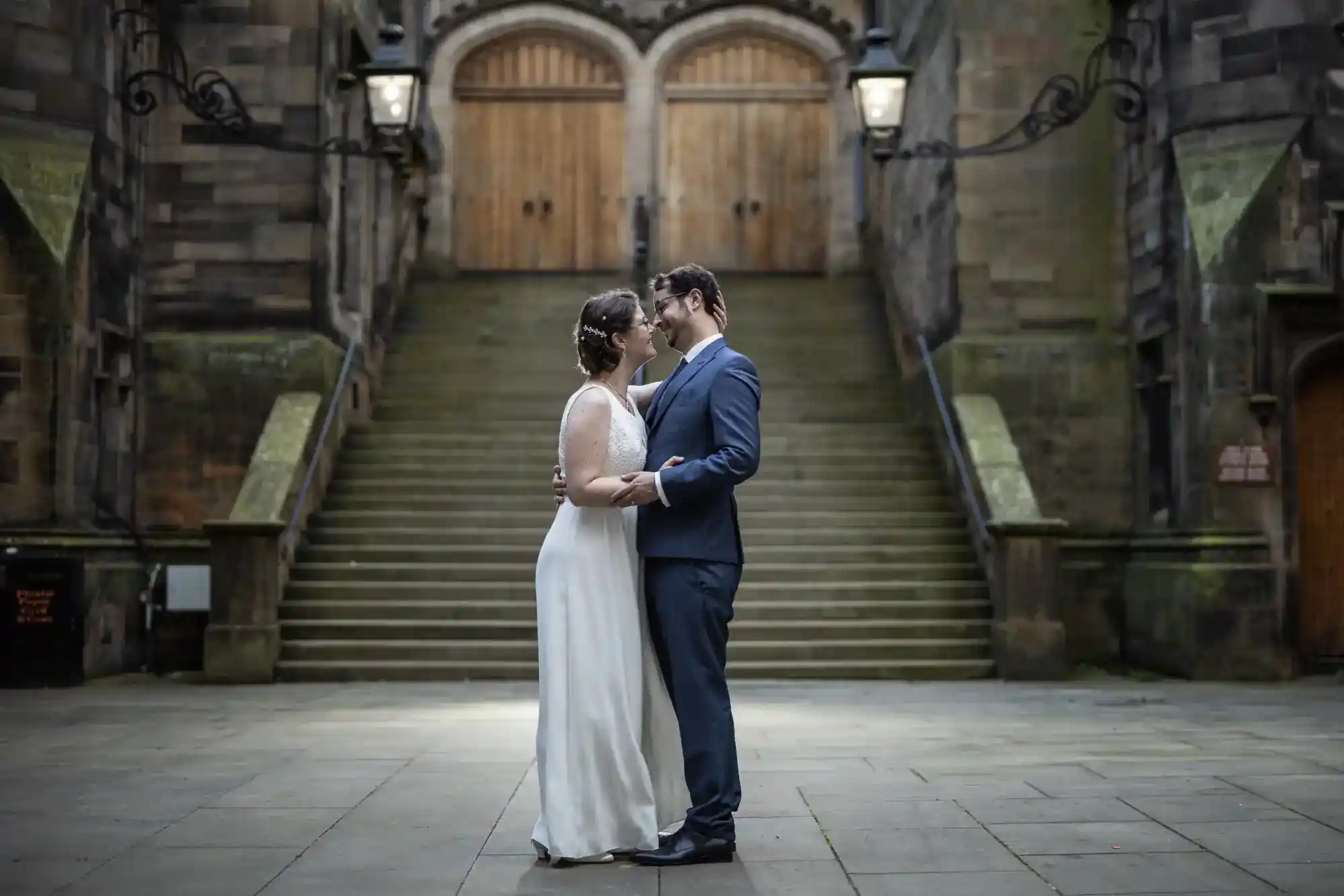 A couple in wedding attire stands close, gazing at each other, in front of wide stone steps and arched wooden doors, under ornate lamps. The setting appears to be an old, grand building with a romantic, historic ambience.
