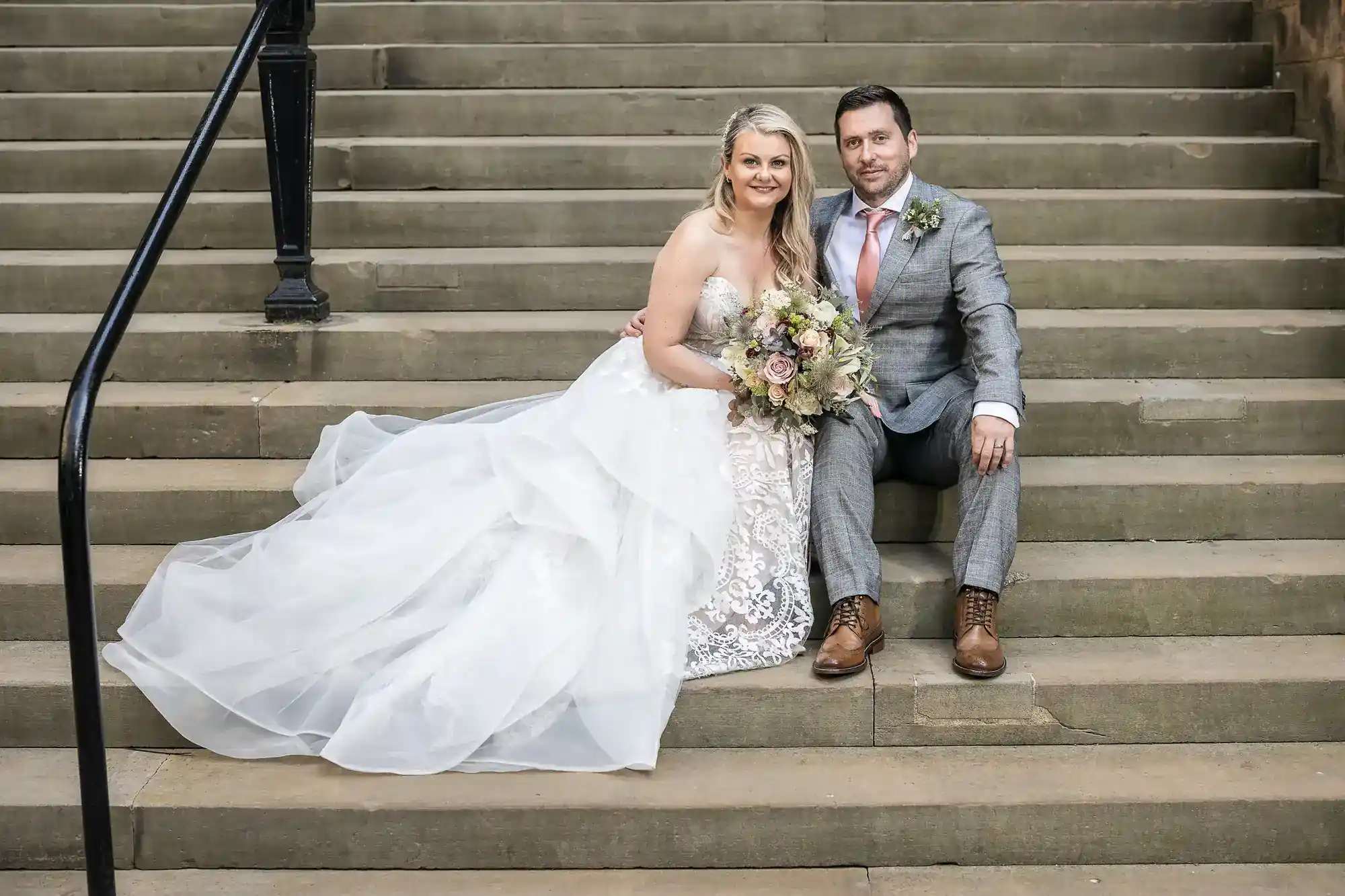A bride in a white lace wedding dress sits beside a groom in a grey suit and pink tie on stone steps. She holds a bouquet of flowers, both smiling at the camera. The bride’s dress flows around them, and black railings frame the scene.
