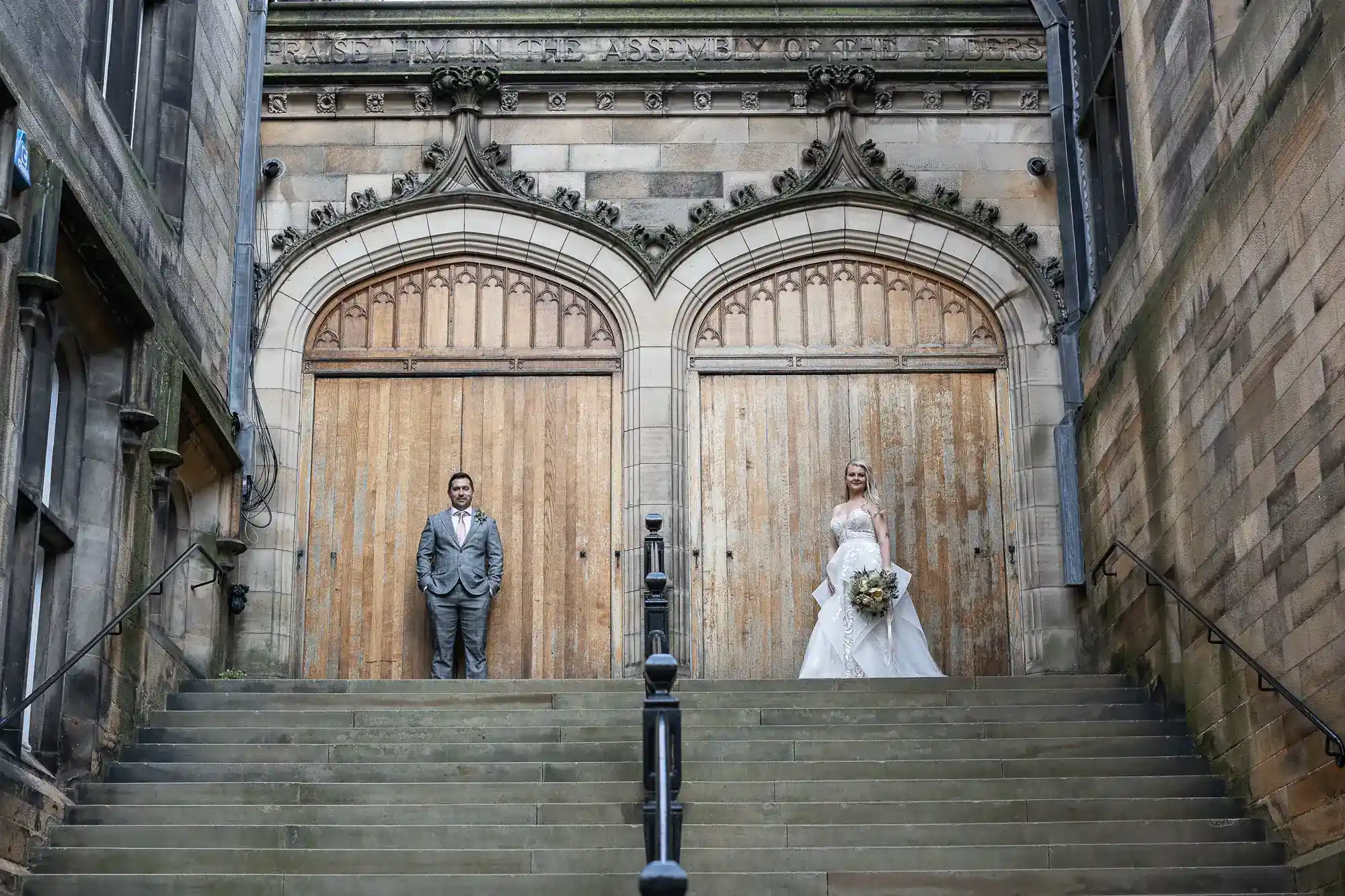 A groom in a grey suit stands on the left, and a bride in a white gown holding a bouquet stands on the right. They pose apart at the top of stone steps, in front of two large wooden doors set in an ornate stone building.