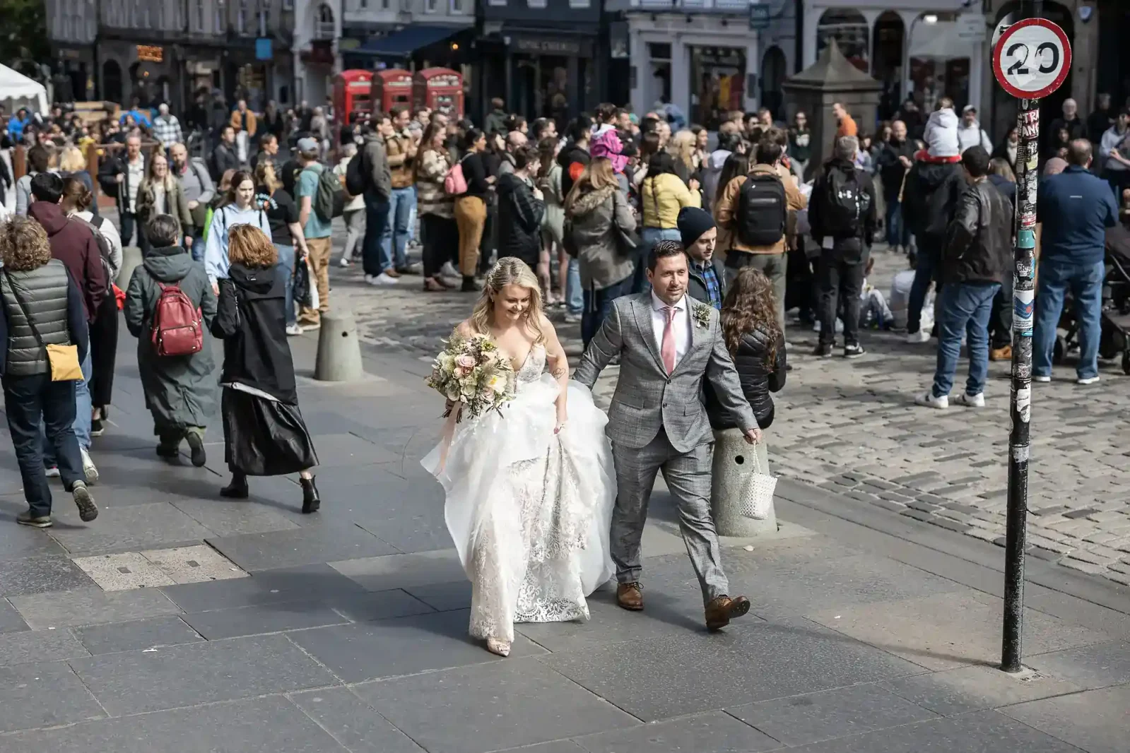 City Chambers newlyweds walking through Festival crowds on the Royal Mile. A bride in a white dress and a groom in a grey suit walk together on a city street. The bride holds a bouquet. Behind them, a large crowd gathers near cobbles, with red telephone boxes and buildings in the background. A 20 mph speed sign is visible.