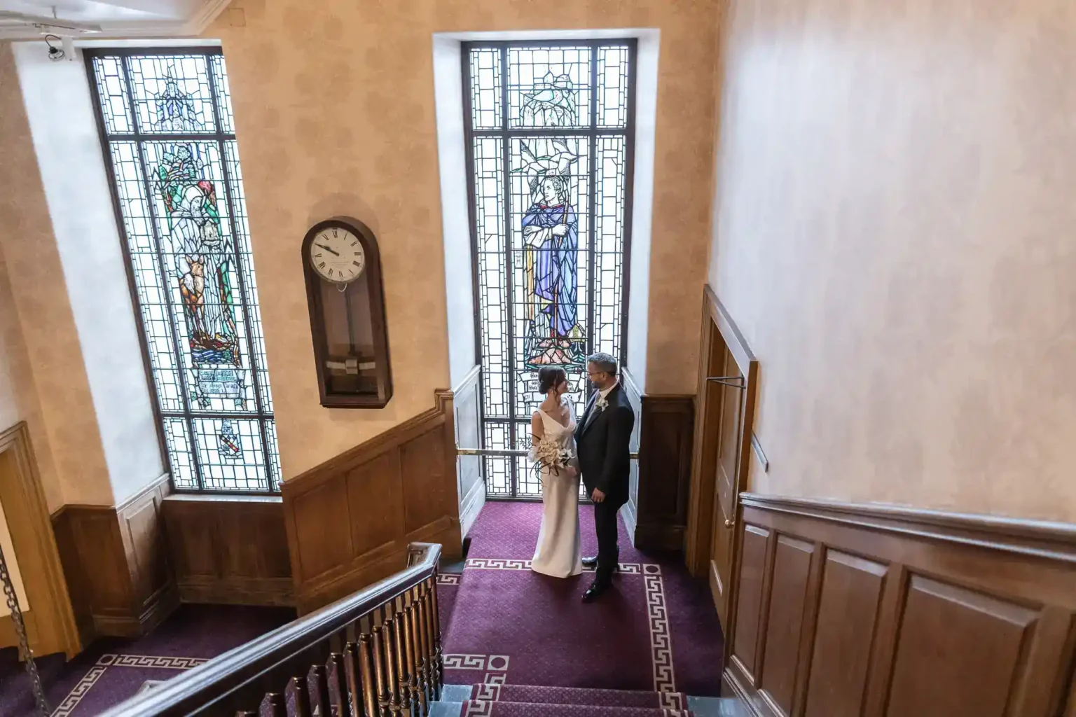 A bride and groom stand together near tall stained glass windows on a stair landing. The room has wood panelling, a red carpet, and a vintage wall clock above the couple. Light streams in from the windows, highlighting their formal wedding attire.