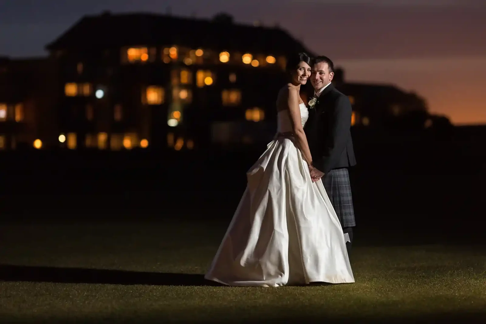 A bride in a white dress and a groom in a dark suit stand together on grass at night, holding hands and smiling. Warm lights from a large building glow softly in the blurred background, creating a romantic atmosphere.