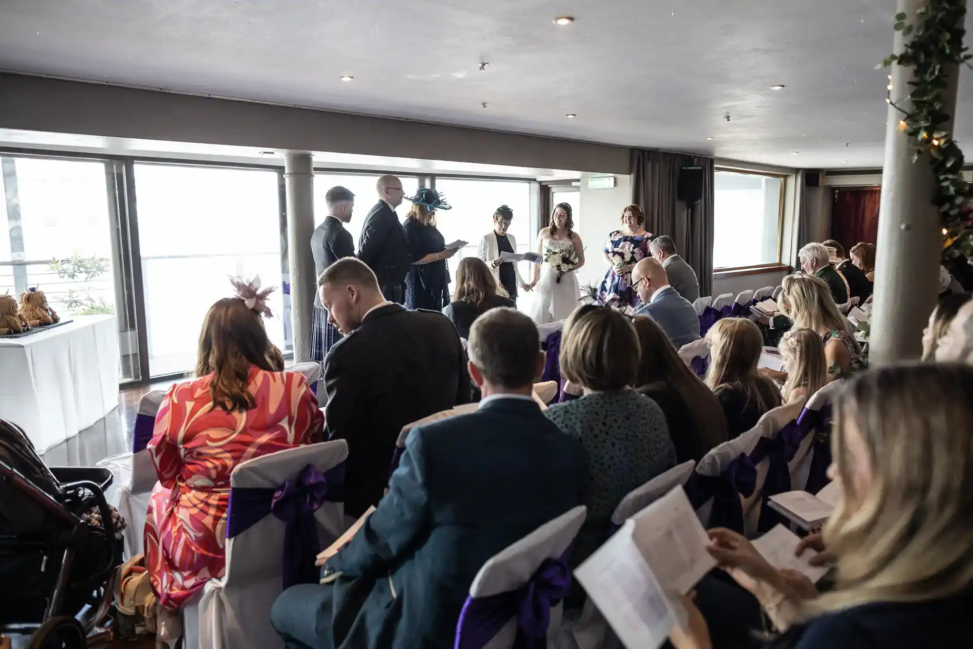 A wedding ceremony takes place indoors with guests seated facing a couple and officiant. The bride in a white dress stands beside the groom and wedding party, while guests, dressed formally, look on. Sunlight shines through large windows behind the wedding party.