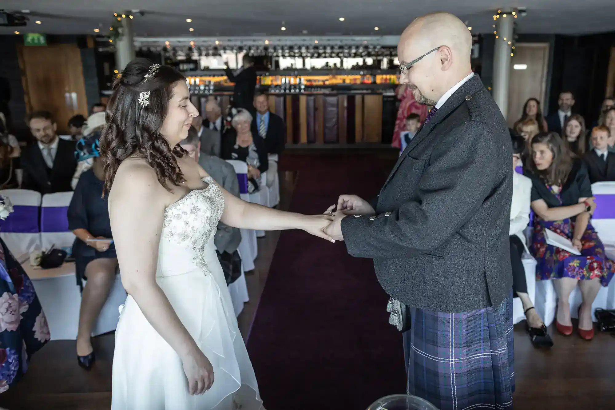 A bride in a white dress smiles as a groom in a kilt places a ring on her finger. Guests are seated in rows behind them, watching the ceremony in a modern, warmly lit venue with a bar at the back.