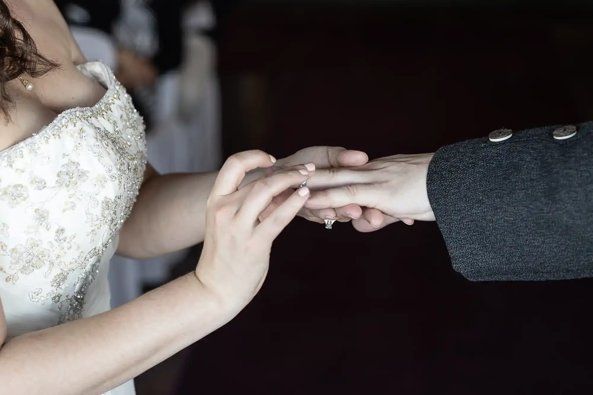 A bride in a white, beaded dress places a ring on her partner’s finger during a wedding ceremony. Only their hands and part of their torsos are visible; the background is dark and out of focus. The moment appears intimate and formal.