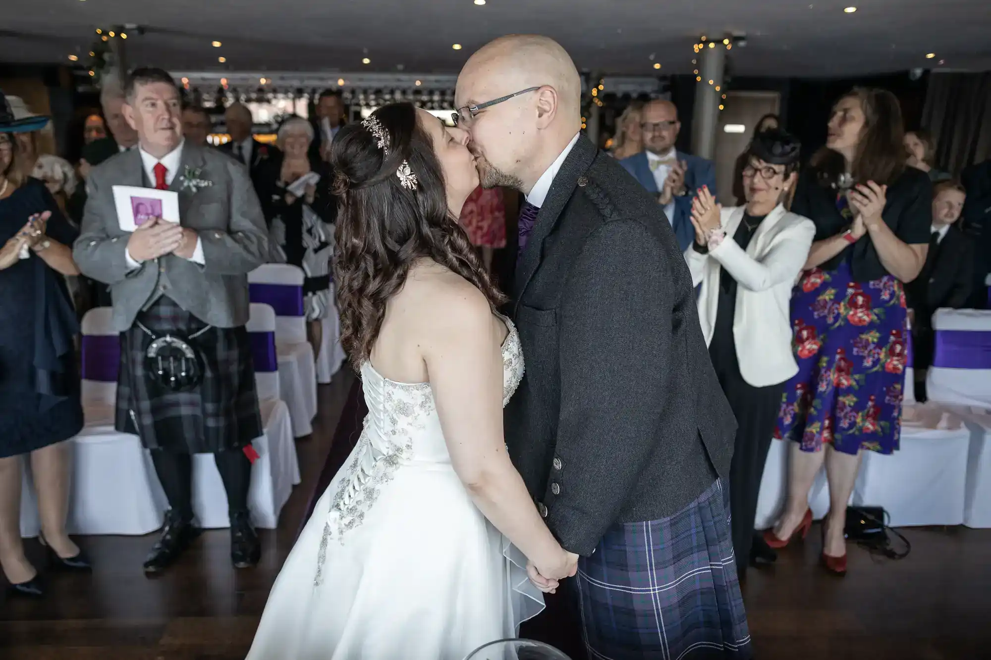 A bride and groom share a kiss at their wedding ceremony, holding hands. The bride wears a white gown, and the groom wears a dark jacket and kilt. Guests, some applauding, watch and smile from decorated chairs in a warmly lit room.