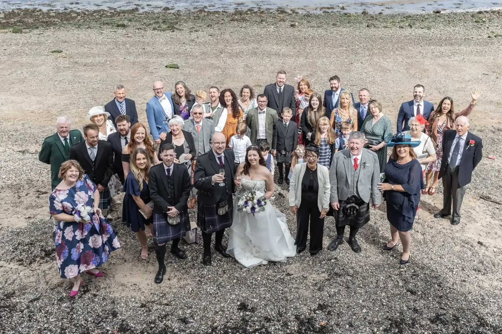 A wedding party stands on a rocky beach, smiling up at the camera. The bride is in white, holding a bouquet, with the groom in a kilt beside her. Surrounding them are well-dressed guests, some in kilts, suits or dresses. The sea and pebbles are in the background.