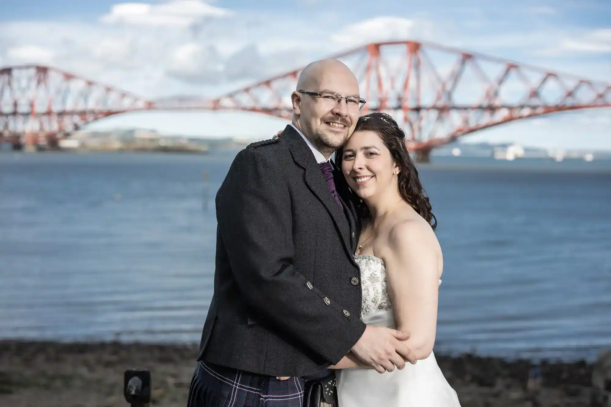 A man in a dark kilt outfit and a woman in a white strapless wedding dress smile and embrace by the water, with a large red steel bridge in the background under a blue sky with clouds.