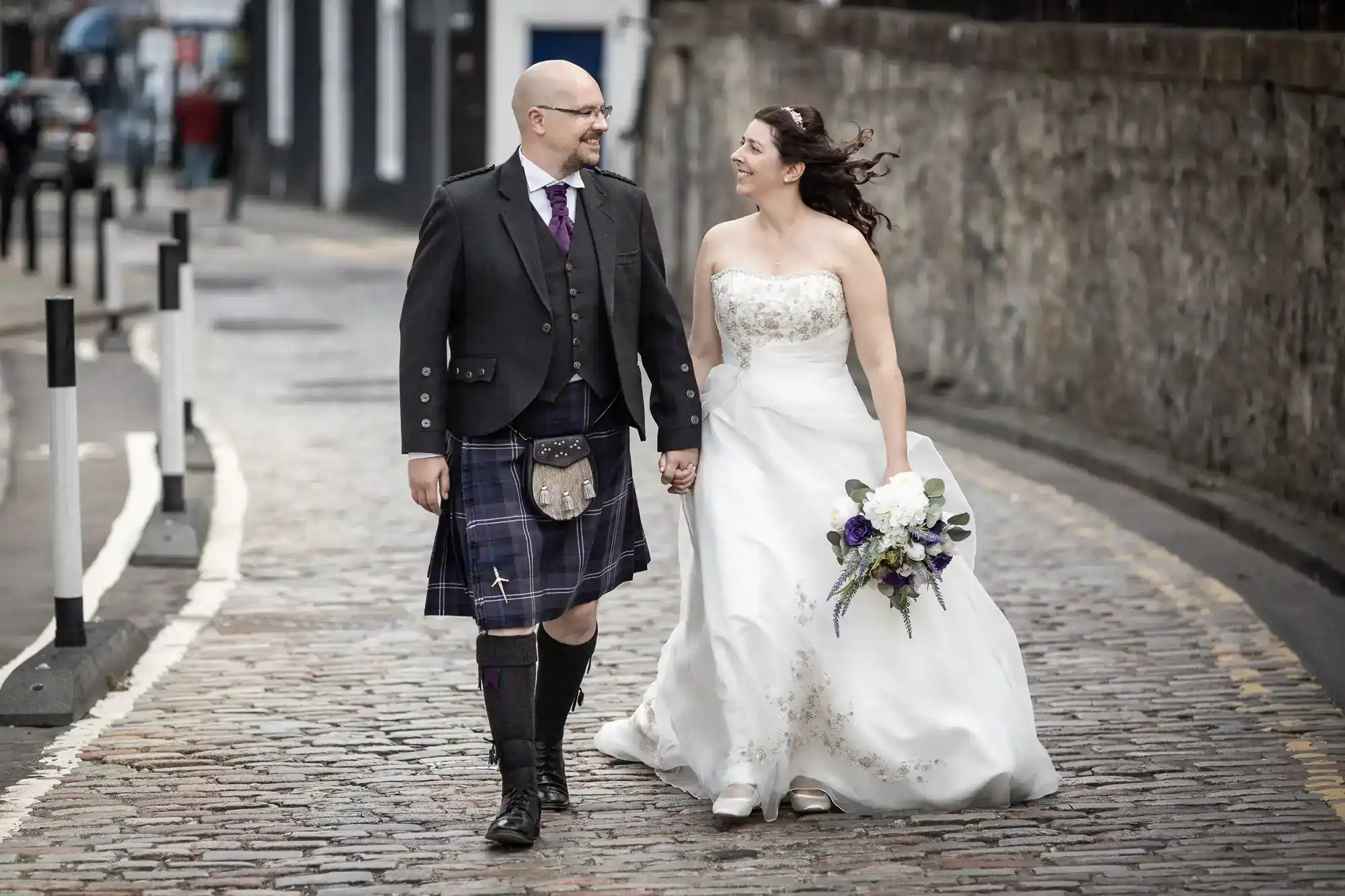 A smiling couple walk hand in hand on a cobbled street. The man wears a traditional Scottish kilt and jacket; the woman, a white wedding dress and holds a bouquet. Both look at each other happily, with a stone wall and blurred buildings behind them.