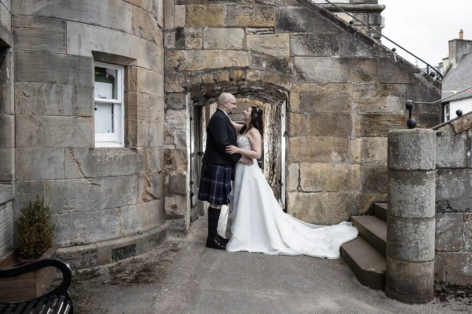 A couple in formal wedding attire embrace and smile at each other under a stone archway. The groom wears a kilt, jacket, and sporran. The bride wears a white gown with a long train. They stand on a stone path next to old stone walls and stairs.