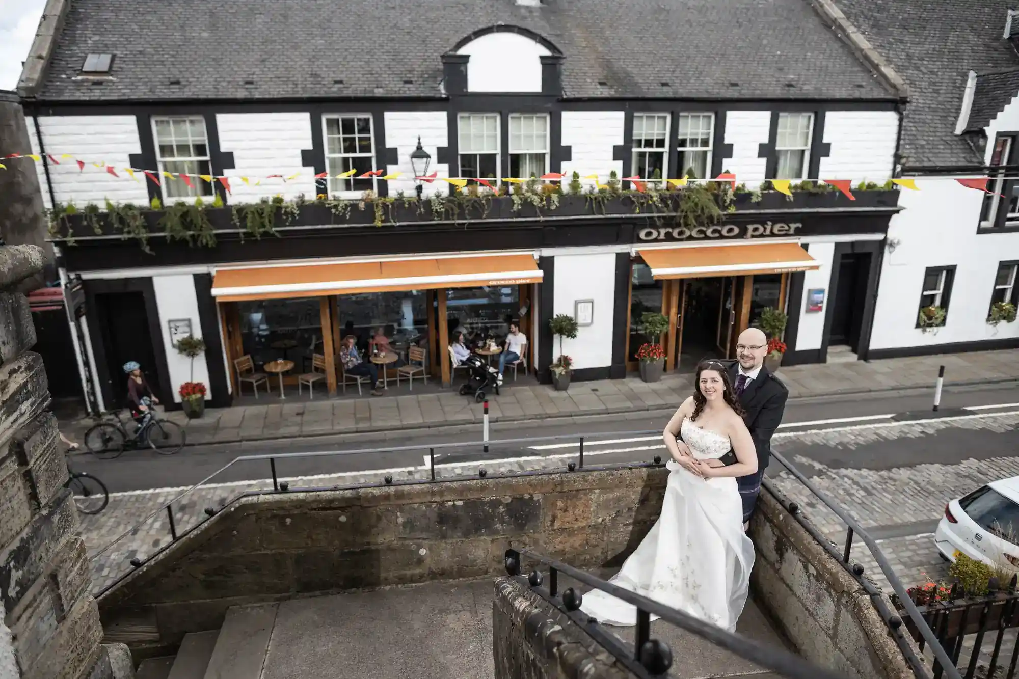 A bride in a white dress and a groom in a dark suit stand on stone steps, smiling and embracing. Behind them is a street, a building with “Orocco Pier” signage, outdoor tables, and festive bunting. The scene appears cheerful and celebratory.