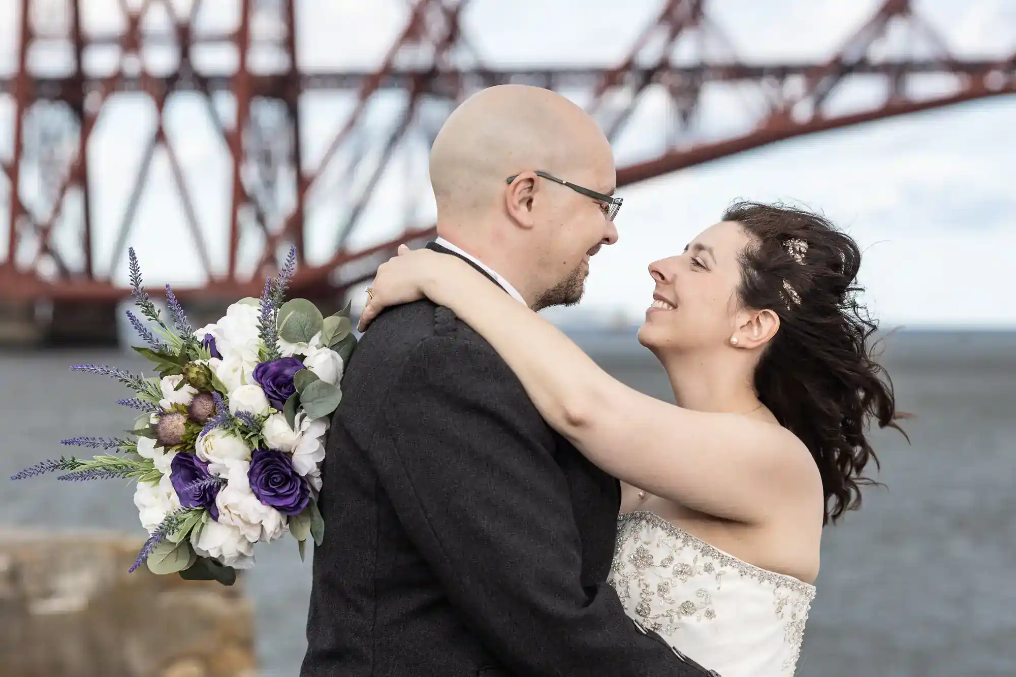 A bride in a white strapless dress embraces a groom in a dark suit. She holds a bunch of white and purple flowers. They smile at each other, standing outdoors near water, with a large red bridge structure blurred in the background.