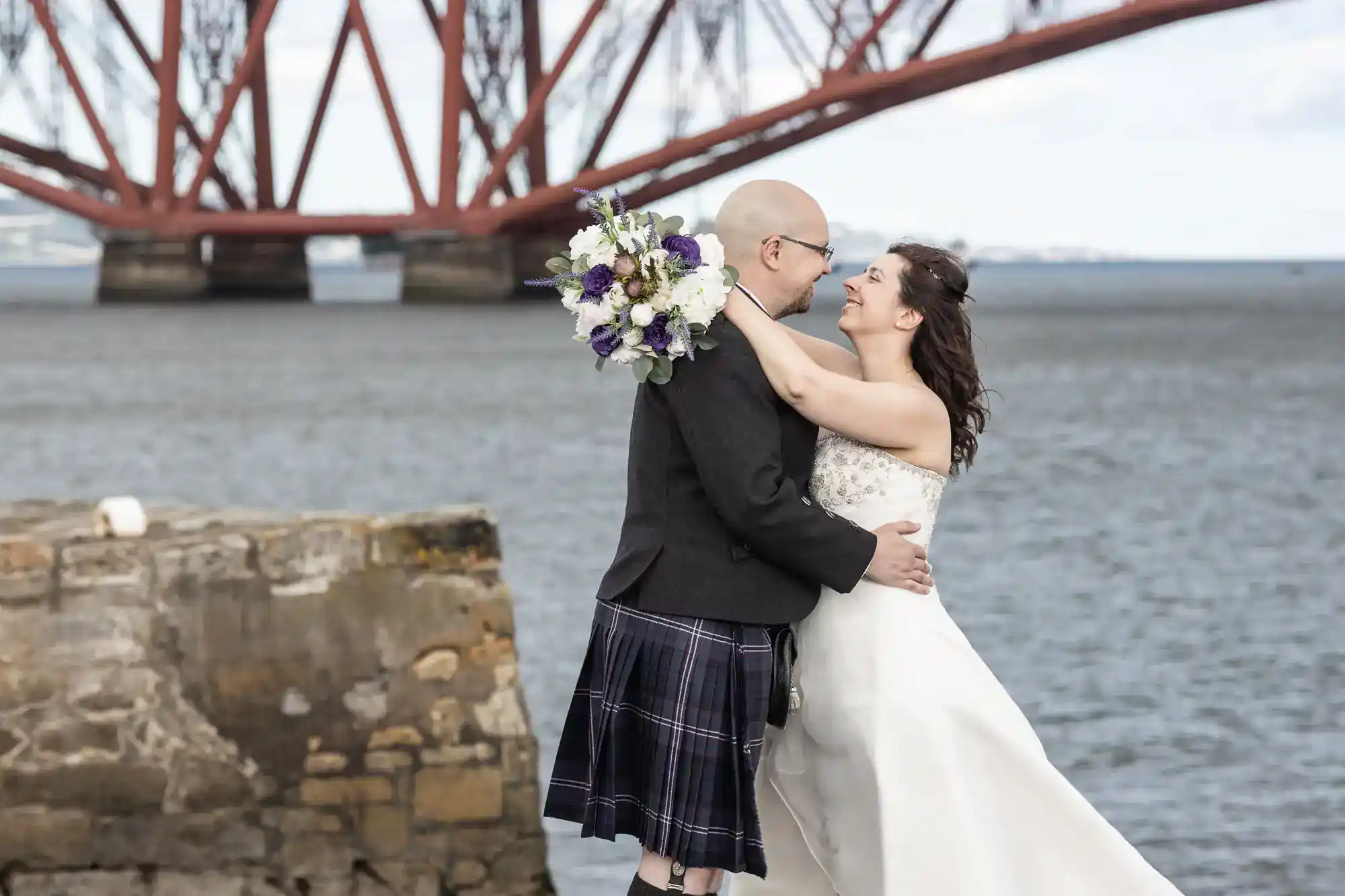 A couple in wedding attire embrace near a stone wall by the water. The groom wears a kilt and glasses, holding purple and white flowers. The bride smiles, arms round his neck. In the background, a large red steel bridge spans the water.