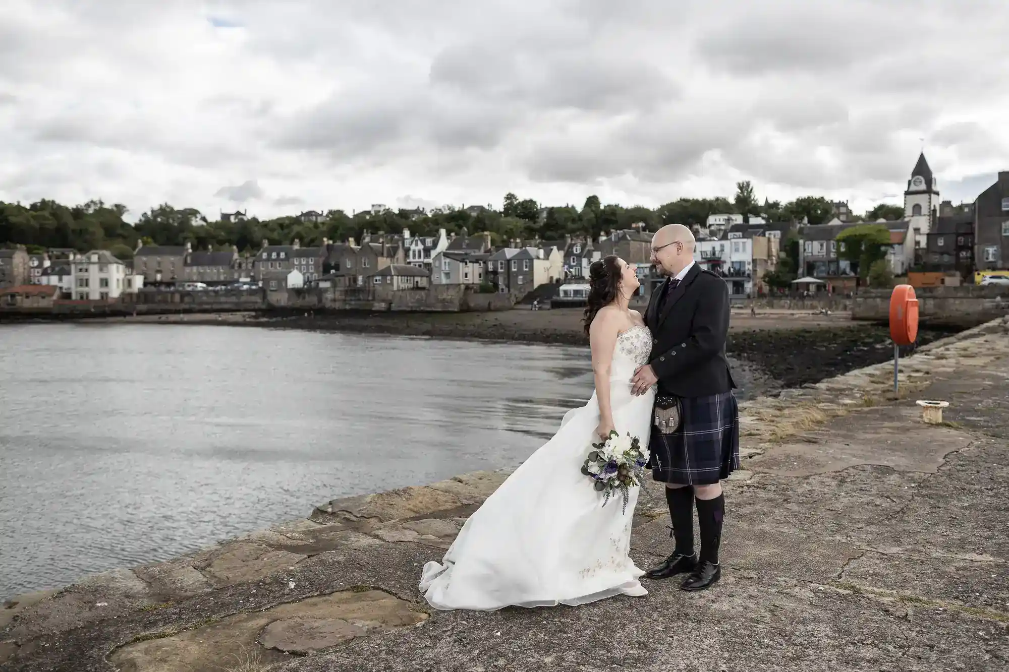 A bride in a white dress and a groom in a kilt stand on a stone quay by calm water, holding each other and smiling. Behind them, there are houses, trees, and a cloudy sky, evoking a small coastal village atmosphere.
