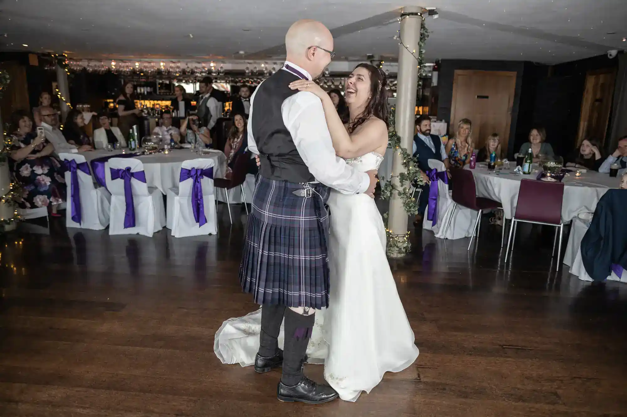 A bride in a white dress and a groom in a kilt share their first dance in a decorated reception hall, surrounded by seated guests watching and smiling. Tables are adorned with white tablecloths and purple sashes, adding a festive touch to the celebration.