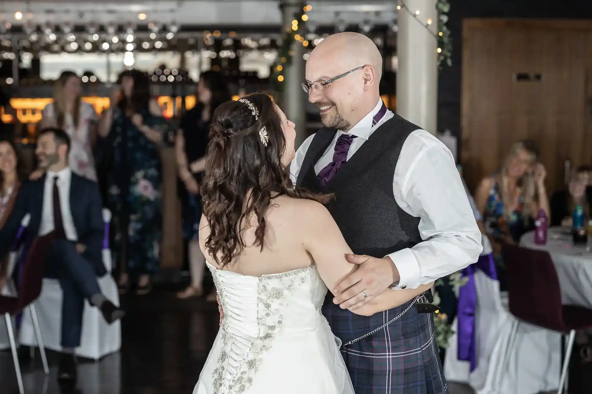 A bride in a white dress and a groom in a dark waistcoat and tartan kilt share a first dance, smiling at each other. Guests sit and watch in the background, with soft lights and decorations visible around the venue.